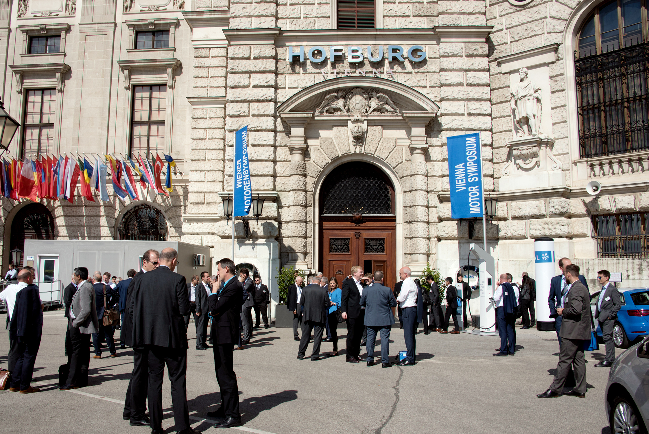 Menschengruppen vor der Hofburg, vor der Fahnen des Motorensymposiums stehen