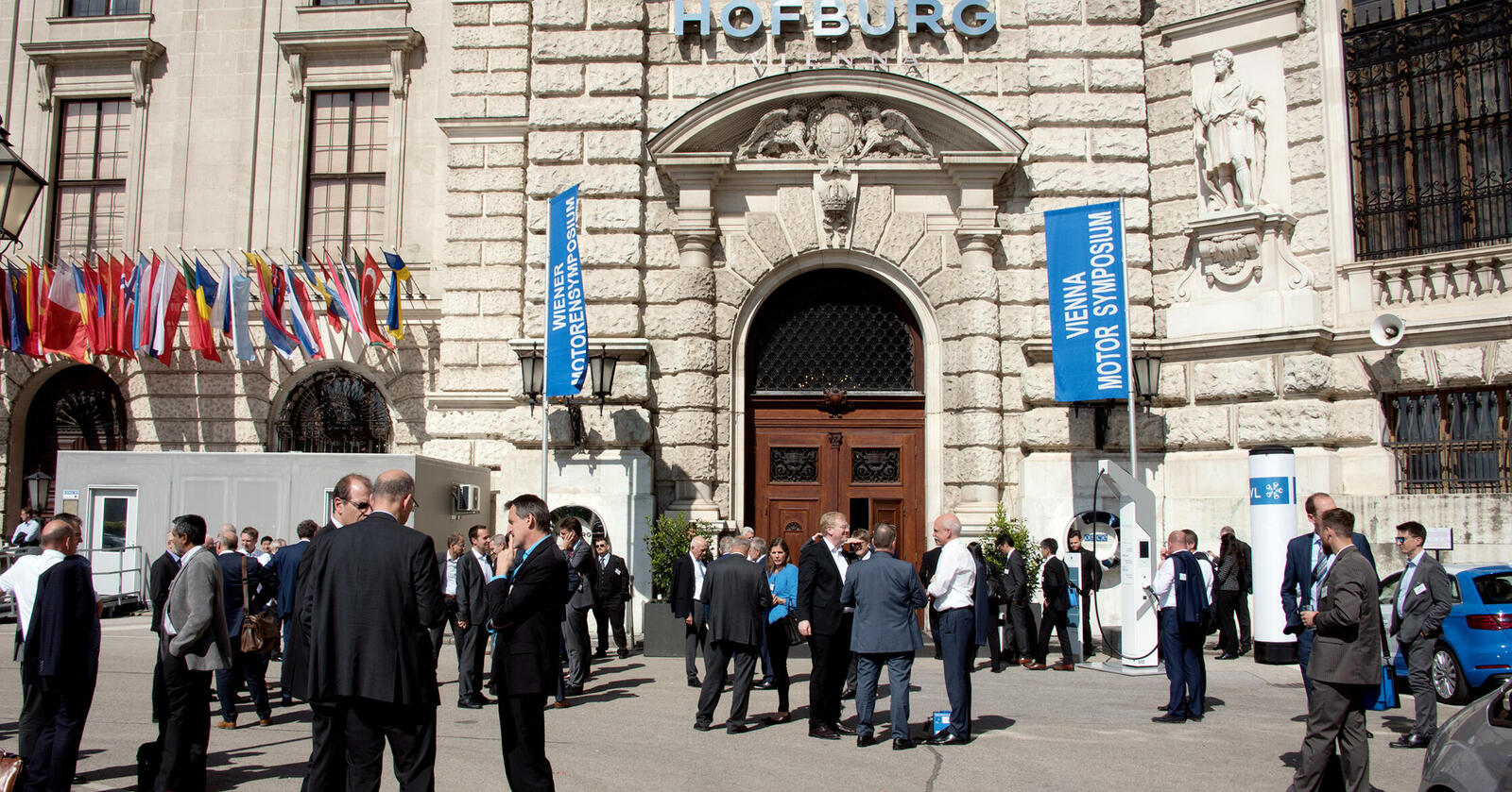Menschengruppen vor der Hofburg, vor der Fahnen des Motorensymposiums stehen