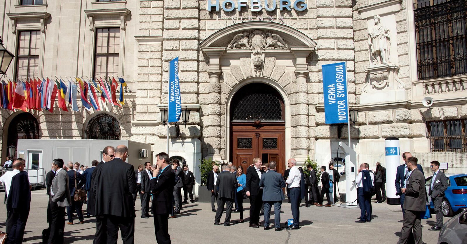 Menschengruppen vor der Hofburg, vor der Fahnen des Motorensymposiums stehen