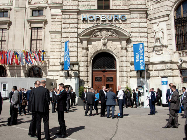 Menschengruppen vor der Hofburg, vor der Fahnen des Motorensymposiums stehen